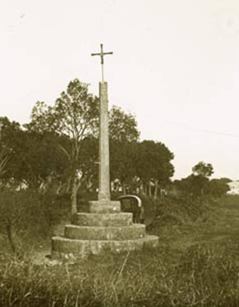 Creu de terme amb uns arbres més enllà i al fons el poble de Palau-Sacosta. 1912