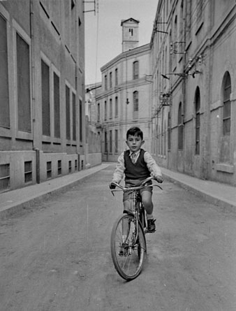 Retrat d'un nen amb bicicleta al carrer Sèquia. A banda i banda, la fàbrica Grober. Al fons, la torre de les Aigües. 1955
