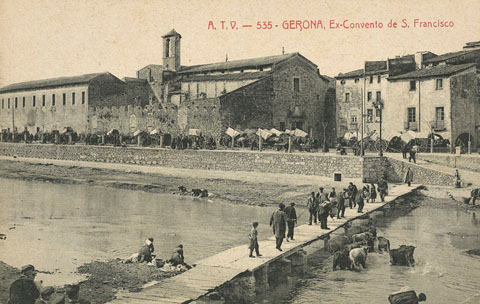 El convent de Sant Francesc de Paula, conegut com els Mínims, i la palanca d'en Vila o de l'Areny vistos de l'altre costat del riu. A l’esquerra, paral·lel al riu, el passeig General Mendoza ple de carruatges durant un dia de mercat i a la dreta, la rambla Pi i Margall. S'observen bugaderes rentant roba al riu. 1905-1907
