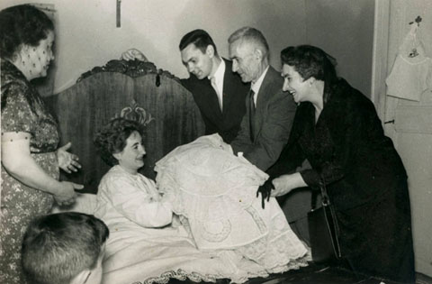 Una mare, al llit, rep el seu fill vestit pel bateig de mans de la llevadora. L'infant és Xavier, el fill de Joan Olivas i Roser Recarens, de Banyoles. 1960
