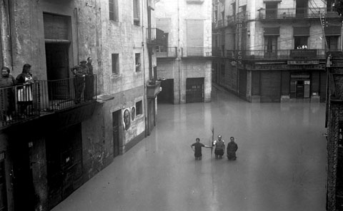 Carrer de la Cort Reial i plaça de l'Oli inundats. A la part central, Daniel Ros Mundet acompanyat de dos nois. A l'esquerra, s'observen cartells del General Franco i de Primo de Rivera. 1940