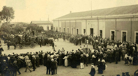 Visita del rei Alfons XIII a la ciutat de Girona. Gent aplegada a la plaça del carril, davant l'estació per rebre el monarca arribat en tren. S'observen diferents carruatges envoltant el vehicle en que viatja el rei i fotògrafs enfilats a sobre d'un carruatge prenent fotografies. 1904