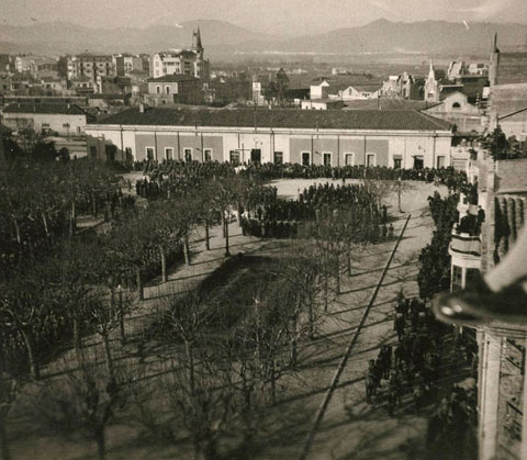 Vista des d'un punt elevat de la carretera Barcelona d'una desfilada militar a la plaça del Carril. Davant l'estació de tren, un altar i gent al voltant. Al fons al centre, l'edifici la Punxa i a la dreta, la farinera Teixidor. 1940-1945