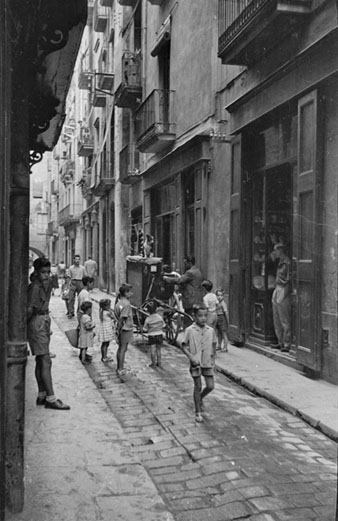Vianants al carrer Mercaders. S'observa un home tocant un organet a la vorera. 1954