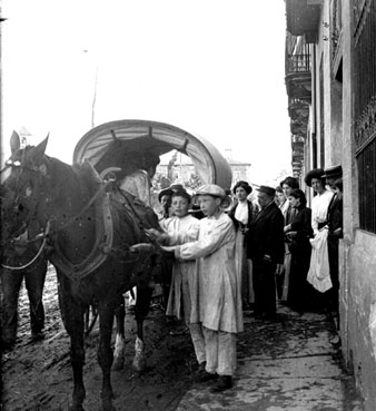 Tartana tirada per un cavall i gent dalt d'una vorera al carrer Camil Mulleras d'Olot. Al fons al centre, l'Hospici. 1910