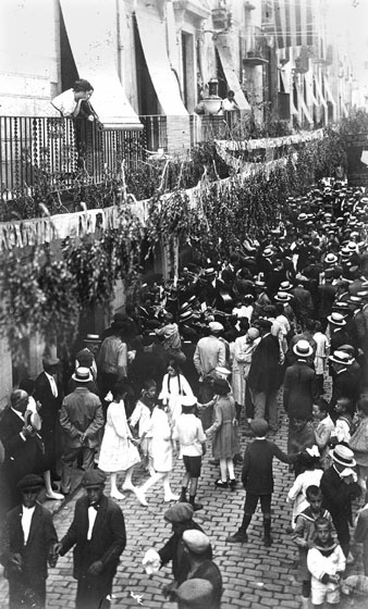 Celebració de la festivitat de Santa Anna al carrer Ballesteries, engalanat amb serrells de paper i branques de boix. Al centre, una cobla tocant sardanes a l'altura de la Casa Masó. 1917