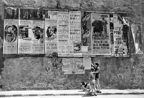 Dos nens amb un gos observant una cartellera d'espectacles i un ban al final del carrer Ballesteries. 1962