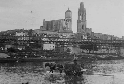 Un carro travessant el riu Ter pel passallís del portal de la Barca cobert d'aigua. En primer terme s'observa el pont del ferrocarril. Al fons l'església de Sant Feliu i la Catedral de Girona. 1920-1925