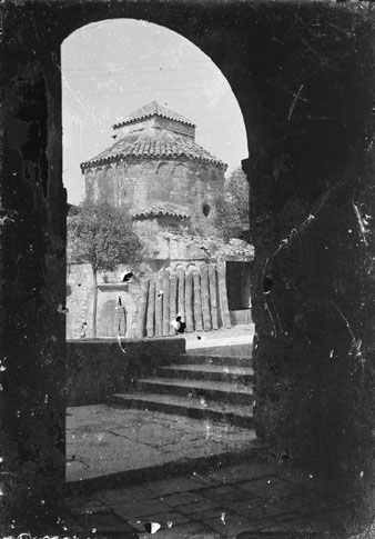Vista de la capella de Sant Nicolau i del carrer de Santa Llúcia des de la porta de l'església de Sant Pere de Galligants. S'observen troncs amuntegats davant de la capella, utilitzada com a magatzem de la fusteria Farreras. 1930-1940